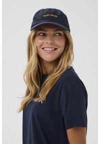 Mornarska kapa za baseball z izvezenim napisom "après tennis" v rumeni barvi, v kombinaciji z mornarsko modro majico s majhnim logotipom na prsih.