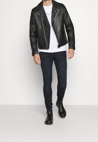 Svart läderbikerjacka med dragkedjor, vit t-shirt, mörka tight jeans och svarta ankelskor, bärs av en stående manlig figur.