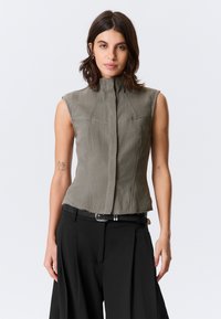 Haut structuré sans manches en vert olive, avec un col montant et des détails de couture à l'avant. Tissu texturé avec un ourlet brut. Ceinture noire en dessous.