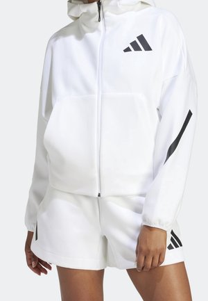 Veste blanche à capuche avec fermeture éclair et shorts assortis, arborant des logos Adidas noirs sur la poitrine, la manche et les shorts, portés par une personne debout.