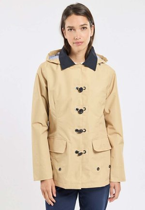 Femme portant une veste à capuche beige avec des boutons à bascule noirs et des poches à rabat, debout devant un fond blanc uni.