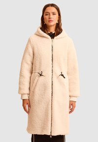 Kremowy, puszysty płaszcz z kapturem, zapinany na suwak z przodu, z regulowanymi troczkami i bocznymi kieszeniami. Posiada prążkowane mankiety i prostą sylwetkę.