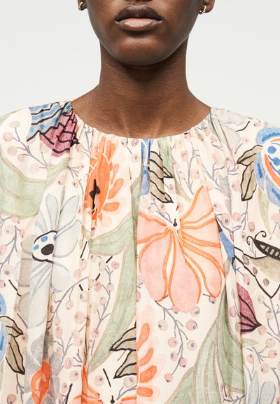 Κοντινό πλάνο ενός ατόμου που φοράει μπλούζα με πολύχρωμο λουλουδάτο σχέδιο σε αποχρώσεις πορτοκαλί, μπλε, πράσινου και ροζ σε ανοιχτό φόντο.