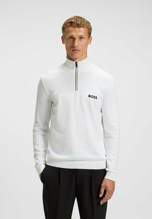 Vit zip-up tröja med texturerat panel, logotyp på vänster bröst, ribbade muddar och fåll. Buren med svarta byxor.
