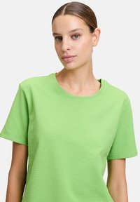 Kwaśnozielona, krótko rękawowa koszulka z teksturowanej tkaniny, z okrągłym dekoltem. Charakteryzuje się luźnym krojem i minimalistycznym wykończeniem.