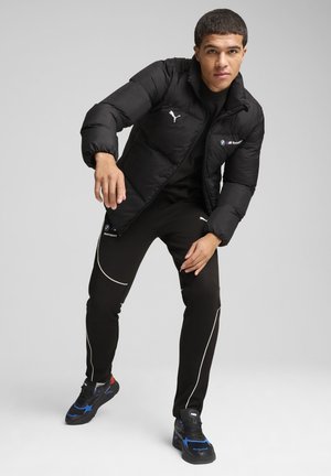 Sort pufferjakke med hvidt Puma-logo, båret over en sort skjorte. Sorte bukser med hvid piping og sorte sneakers med blå accenter.
