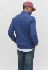 Blå våffelstickad pullover med hög krage, har ett ribbat avslut. Kombinerad med en ljusblå skjorta med krage och mörka jeans. Kappen är rosa.