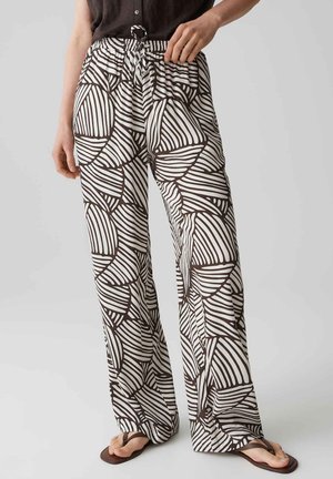 Weit geschnittene weiße Hose mit schwarzen abstrakten gebogenen Linienmustern, getragen mit braunen Flip-Flops und einem dunklen Oberteil, Hände ruhen nahe der Taille.