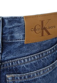 Jeans in denim con un lavaggio azzurro chiaro, caratterizzati da un'etichetta in pelle beige sulla vita con il logo "Calvin Klein Jeans" impresso, e dettagli di cucitura visibili.