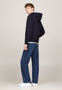 Sudadera azul marina con capucha ajustable, combinada con jeans de mezclilla oscuros de corte holgado y zapatillas blancas; diseño simple con puños y dobladillo acanalados.