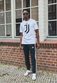 Hvid t-shirt med sort Juventus-logo, parret med sorte træningsbukser med hvide striber og Adidas-logo, samt hvide sneakers med røde accenter.