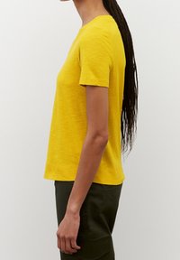 Gul kortærmet t-shirt med rund hals, struktureret stof og lige pasform. Båret med mørkegrønne bukser, vist fra siden.