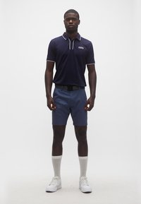 Άνδρας στέκεται προς τα εμπρός φορώντας μπλε ναυτικό πόλο πουκάμισο, μπλε σορτς με μαύρη ζώνη, λευκές κάλτσες μέσης σύστασης και λευκά αθλητικά παπούτσια σε λευκό φόντο.