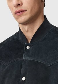 Giacca in suede nera con finitura testurizzata, colletto a coste e bottoni a scatto argento. Presenta dettagli di cucitura a yoke curvo.