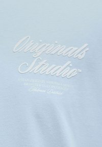 Svetlomodré bavlnené tričko s vyvýšenou bielou výšivkou s nápisom "Originals Studio" a ďalším textom nižšie. Hladká textúra, ležérny dizajn.