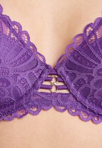 Reggiseno in pizzo viola con motivi floreali, bordi ondulati e un accento in metallo dorato al centro con dettagli delle spalline.