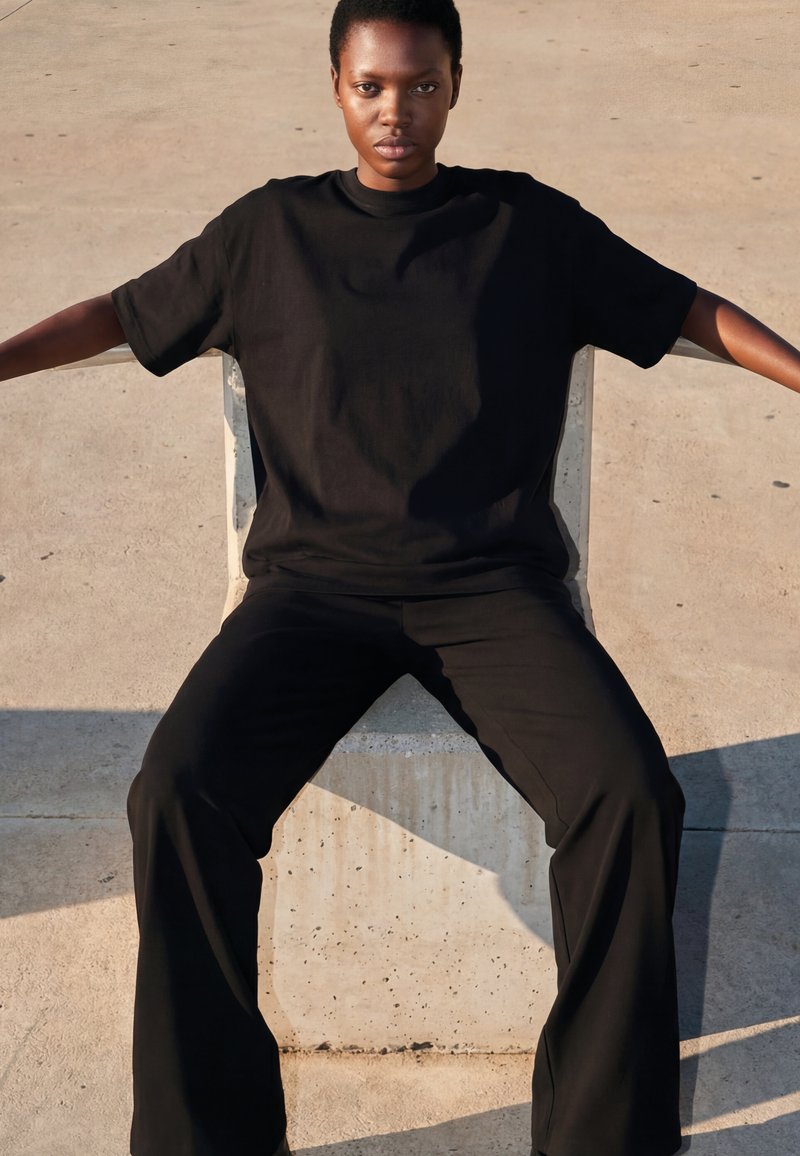 Mladý člověk v uvolněné pozici, oblečený v černém tričku a kalhotách, sedící venku na betonovém povrchu s rozpaženýma rukama.