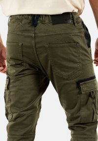 Pantalons cargo vert olive avec une texture douce, taille élastique, de multiples poches et des détails de couture subtils. Présente des accents en métal noir.