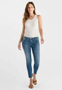 Witte mouwloze top, blauwe spijkerbroek met een slanke pasvorm en bruine sandalen. De spijkerbroek heeft een klassieke knoop- en ritssluiting, met een lichte wassing en lichte vervaging.