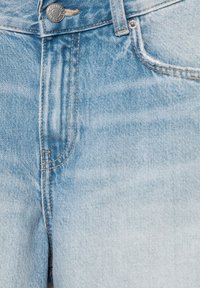 Jean en denim bleu clair en gros plan montrant le bouton avant, la zone de la fermeture éclair et la poche avec des détails de couture visibles.