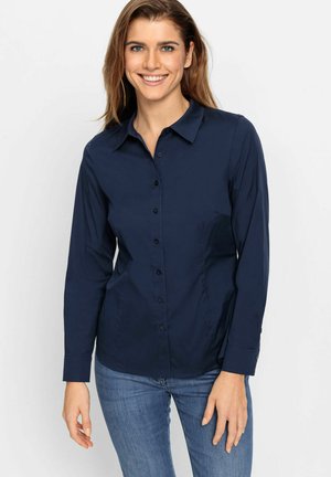 Glimlachende vrouw met lichtbruin haar, gekleed in een donkerblauw blouse met knopen en blauwe spijkerbroek, staand tegen een effen witte achtergrond.