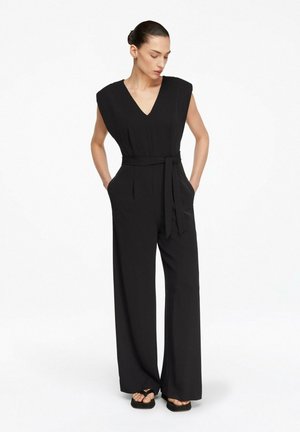 Kobieta ubrana w czarny, bezrękawnikowy kombinezon z szerokimi nogawkami, z dekoltem w serek i paskiem w talii, stoi z rękami w kieszeniach, patrząc w dół.