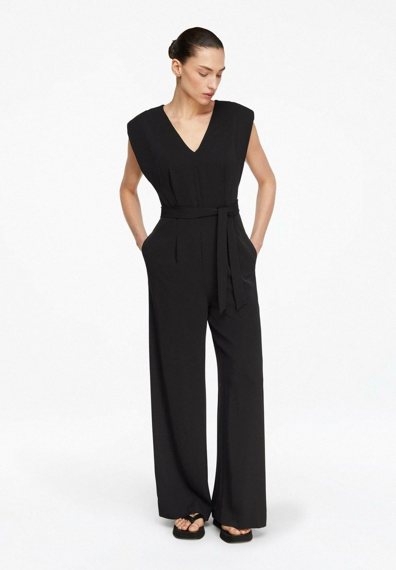 Kobieta ubrana w czarny, bezrękawnikowy kombinezon z szerokimi nogawkami, z dekoltem w serek i paskiem w talii, stoi z rękami w kieszeniach, patrząc w dół.