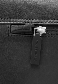 Angolo di borsa in pelle nera con una cerniera argentata, superficie liscia e una targhetta rettangolare nera con la scritta "STRELLSON" in rilievo.