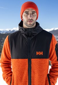 Flísová bunda s černou horní částí a oranžovou texturovanou dolní částí, se zapínáním na přední zip a kapucí. Obsahuje detail s logem na hrudi.