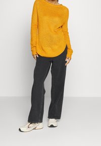 Persona che indossa un maglione giallo senape a maglia larga, jeans neri svasati e sbiaditi, e sneaker bianche con dettagli marroni.