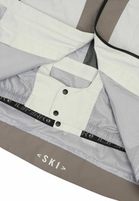 Innenansicht einer Skijacke in Grau und Weiß, mit Druckknopfverschlüssen, elastischem Bund und einer Netztasche. Auffälliges Logo am unteren Ende aufgedruckt.