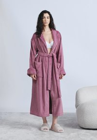 Γυναίκα με μακριά, σγουρά μαλλιά που φοράει μωβ ρόμπα πάνω από λευκό σουτιέν και χνουδωτές παντόφλες, στέκεται σε γκρι χαλί κοντά σε μια καμπύλη λευκή καρέκλα.