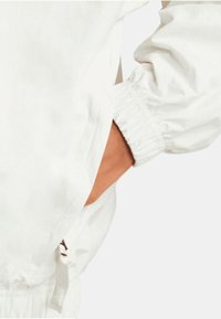 Veste blanche avec des poignets élastiques et une poche sur le côté. Le tissu semble lisse avec une finition mate.