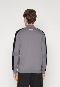 Mann mit grauem und schwarzem Langarmpullover mit "BOSS" auf dem oberen Rücken, kombiniert mit schwarzen Hosen, von hinten vor einem weißen Hintergrund gezeigt.