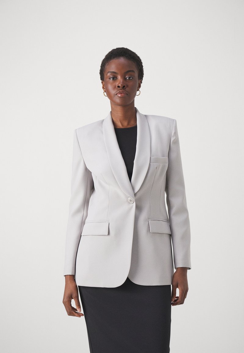 Blazer gris claro con un diseño estructurado, solapas con muesca, cierre de un solo botón y dos bolsillos frontales, usado sobre una blusa negra.