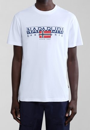 Hombre con camiseta blanca de Napapijri con texto azul marino y gráfico de la bandera noruega, combinado con pantalones azul oscuro sobre fondo gris.
