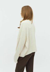 Pull en maille côtelée crémeux avec une coupe décontractée, des épaules tombantes et une texture douce, associé à un pantalon brun foncé.