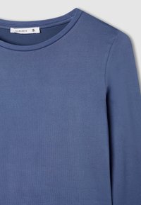 Niebieska bluzka z długim rękawem, wykonana z gładkiego materiału, z okrągłym dekoltem, posiadająca prosty fason oraz małą metkę wskazującą rozmiar.