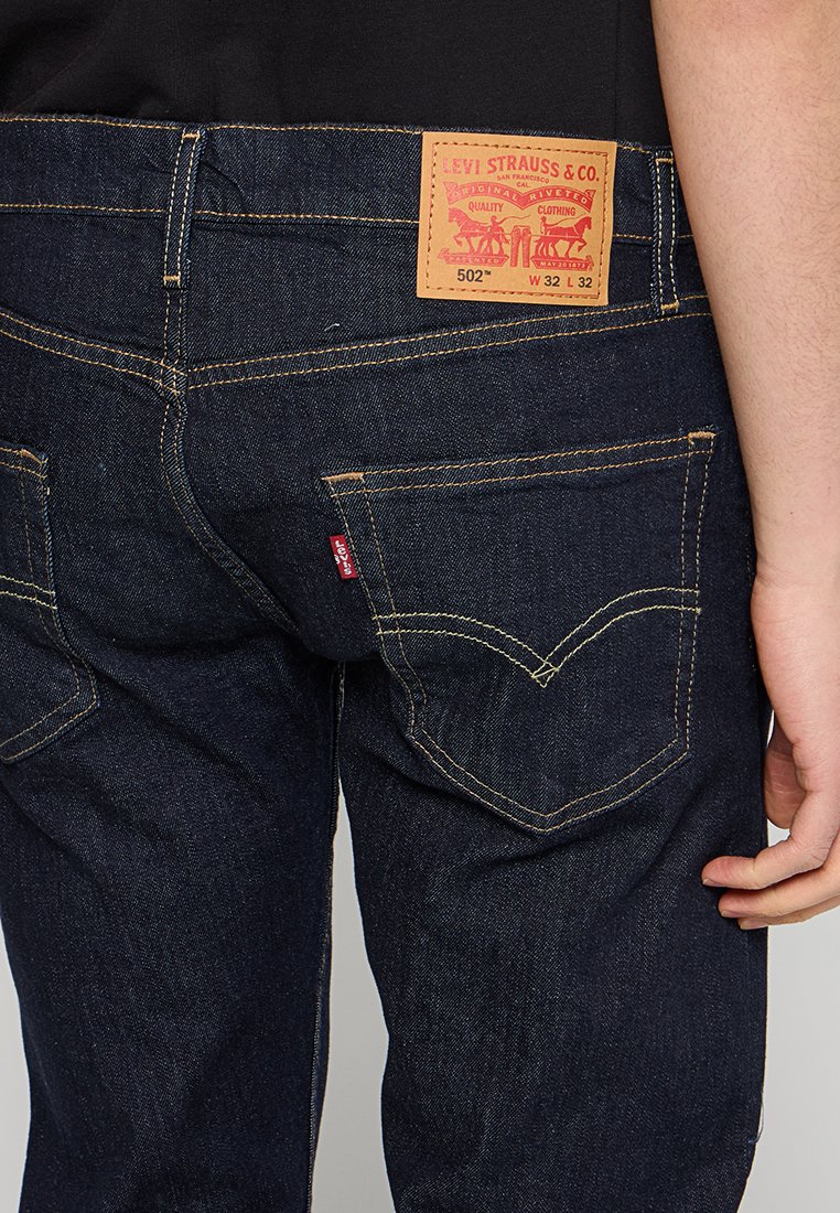 Jean en denim bleu foncé avec une patch en cuir à l'arrière présentant du texte et des graphiques rouges, des coutures jaunes et deux poches arrière avec des coutures caractéristiques.