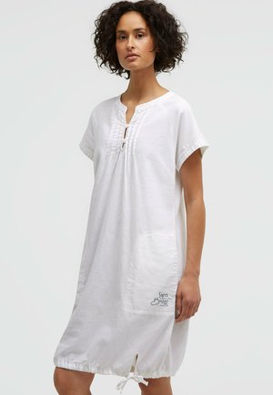 Woman with curly hair wearing a loose white knee-length dress with short sleeves and a front pocket with text print.