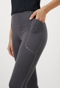 Szare legginsy z wysokim stanem, wykonane z elastycznego materiału, z boczną kieszenią i odblaskowym wykończeniem wzdłuż krawędzi kieszeni.