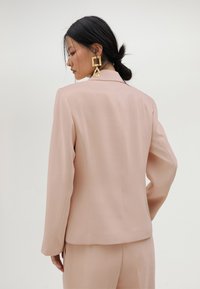 Ružičasti krojeni sako s dugim rukavima, strukturiranim ramenima i glatkom završnom obradom. Ima jedan stražnji prorez za lakše kretanje.