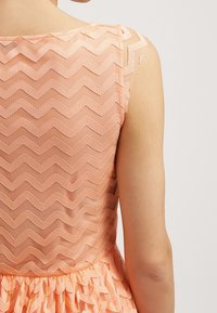 Robe couleur pêche avec un tissu texturé en zigzag, détails transparents aux épaules, jupe plissée et corsage ajusté.