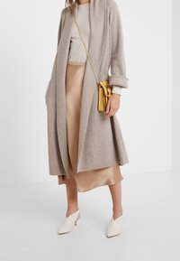 Beige ribbed pitkä neuletakki, kermajäykät yläosat, satiininen beige hame ja valkoiset avonaiset mules-kengät. Kultaista olkalaukkua lisää väripiristystä.