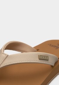 Κοντινό πλάνο λουριού σανδαλιού μπεζ υφής με μεταλλική πλακέτα μάρκας REEF σε ταμπά πάτο, μπροστά σε φωτεινό φόντο.