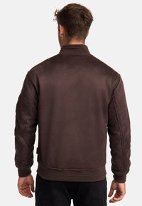 Chaqueta bomber marrón con textura suave, puños y dobladillo acanalados, que presenta un respaldo fruncido y detalles de bolsillo lateral.