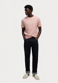 Jasnoróżowa bawełniana koszulka, ciemne dżinsy i szare trampki. Model stoi z rękami w kieszeniach, prezentując swobodny krój i kolory.