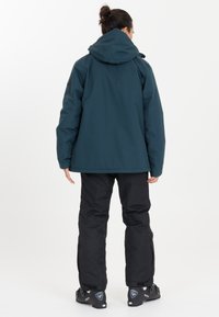 Teal waterdichte jas met een aangehechte capuchon, gecombineerd met zwarte geïsoleerde broek en zwarte sneeuwlaarzen met versterkte zolen en accenten.