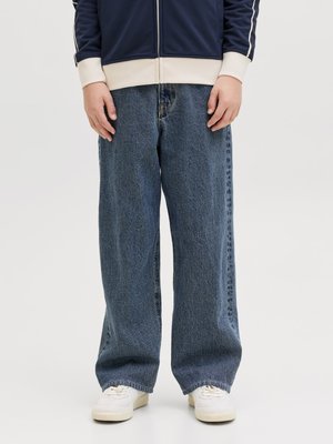 Person iført løstsiddende blå jeans, hvide sneakers og en navy jakke med beige manchetter, stående mod en ensfarvet baggrund.