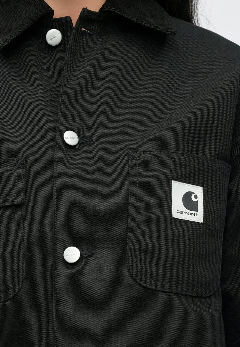 Chaqueta negra Carhartt con botones de presión metálicos y parche con logo cuadrado en el bolsillo izquierdo del pecho, llevada por una persona.